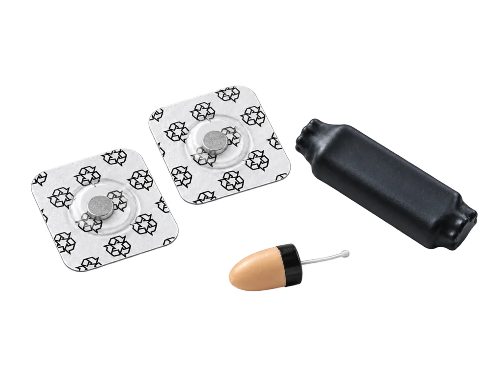 Editorial render of a beige miniature earpiece, a black module and two button-cell battery packs.